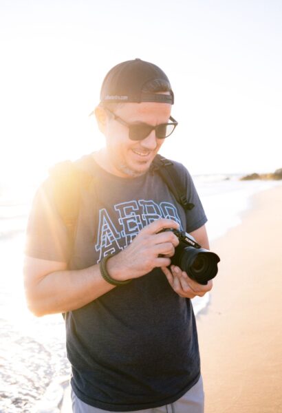 A man wearing sunglasses and a backwards cap stands on a sunny beach, looking down and adjusting a camera in his hands.